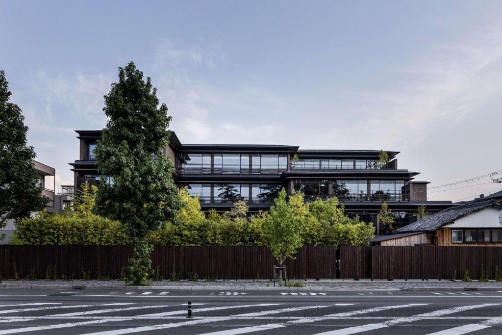 Modern multi-story building with large glass windows, partially hidden behind tall green trees and a wooden fence. A crosswalk and paved street are visible in the foreground under a clear sky.