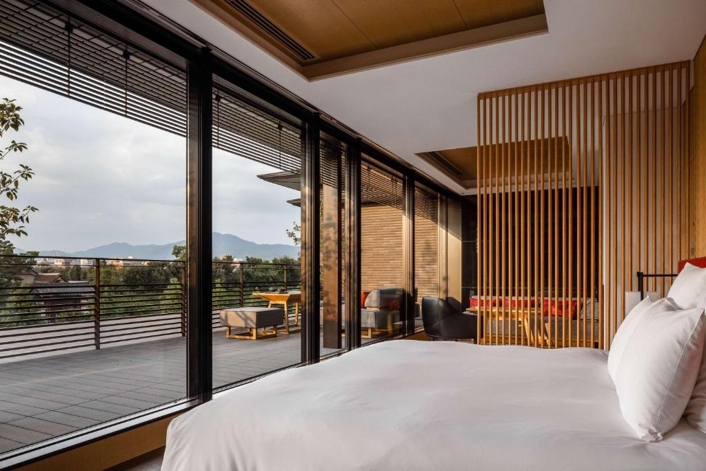 Modern bedroom with white bedding, floor-to-ceiling windows, wooden slat divider, and a spacious balcony featuring outdoor seating; mountain and city views visible in the background.