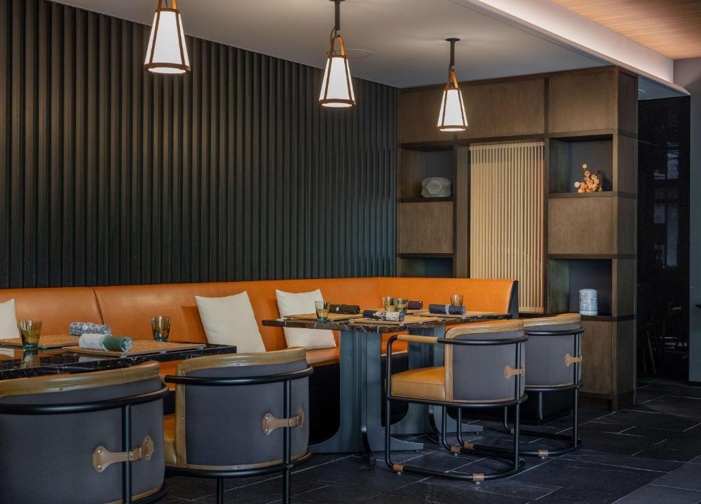 A modern restaurant interior with brown leather seating, black tables, cushioned chairs, pendant lights, and a wall with vertical panels and open shelving displaying decor items.