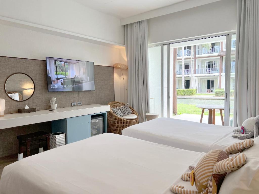 Bright hotel room with two neatly made beds, a wicker chair by a large window, a desk with a round mirror, a wall-mounted TV, and a view of a green lawn and resort buildings outside.