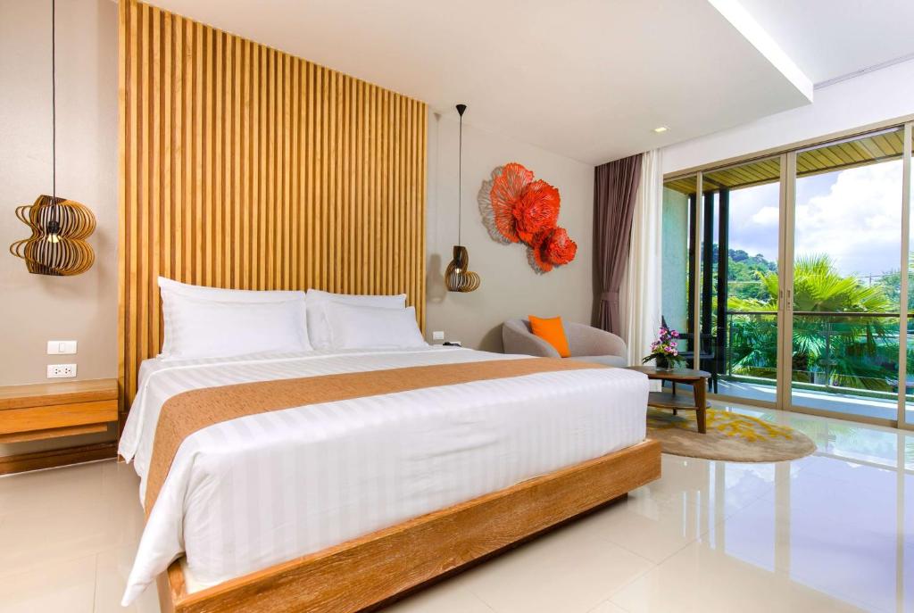 Modern hotel room with a large bed, wooden accents, stylish lighting, decorative orange wall art, a gray chair with an orange pillow, and floor-to-ceiling windows overlooking greenery and a blue sky.