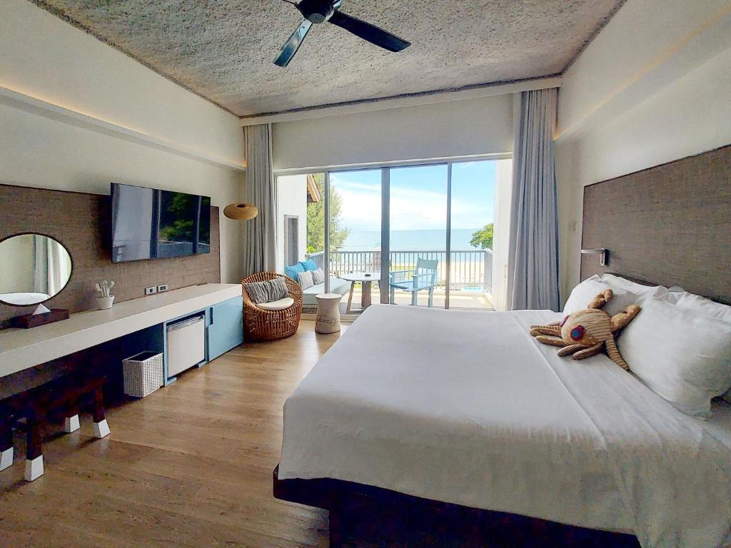 Bright hotel room with a large bed, pillows, and two teddy bears; desk and TV on one side, a wicker chair near glass doors that open to a balcony with ocean view. Light wood floors and ceiling fan complete the airy space.