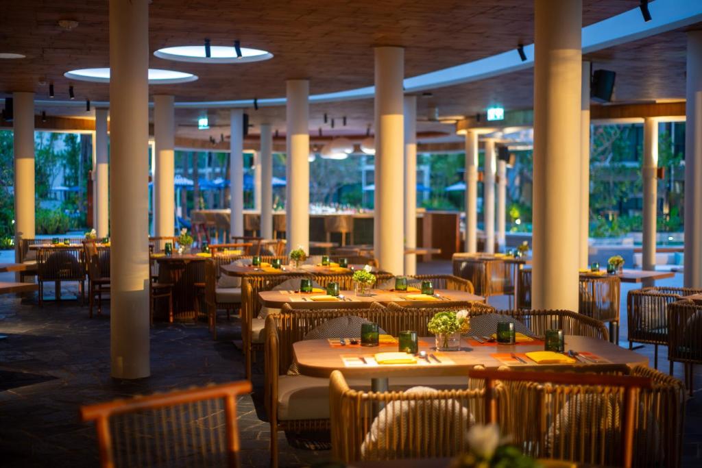 A modern, warmly lit restaurant with neatly arranged tables and chairs, each table decorated with small flower vases. Large windows reveal greenery outside, and round ceiling lights enhance the cozy, inviting atmosphere.