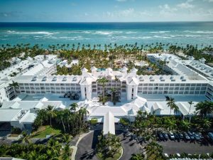 Aerial view of a large white beachfront resort surrounded by palm trees, with the ocean and waves in the background and a parking lot with cars in the foreground.