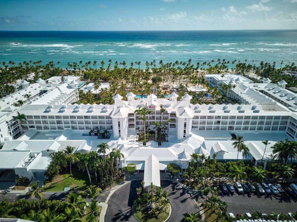 Aerial view of a large white beachfront resort surrounded by palm trees, with the ocean and waves in the background and a parking lot with cars in the foreground.
