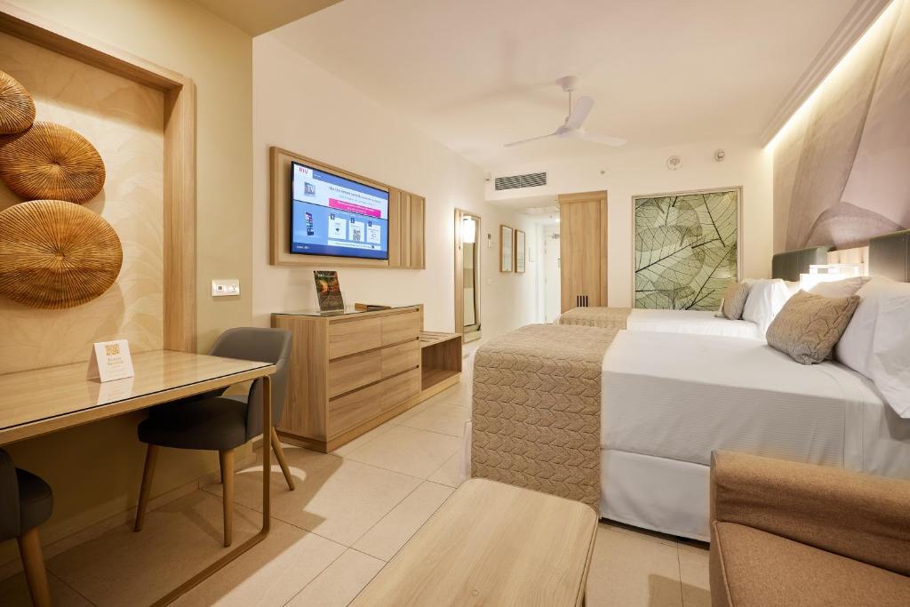 Modern hotel room with two double beds, a wall-mounted TV, a small dining table with chairs, a wooden dresser, and neutral decor with beige tones and nature-inspired artwork on the wall.