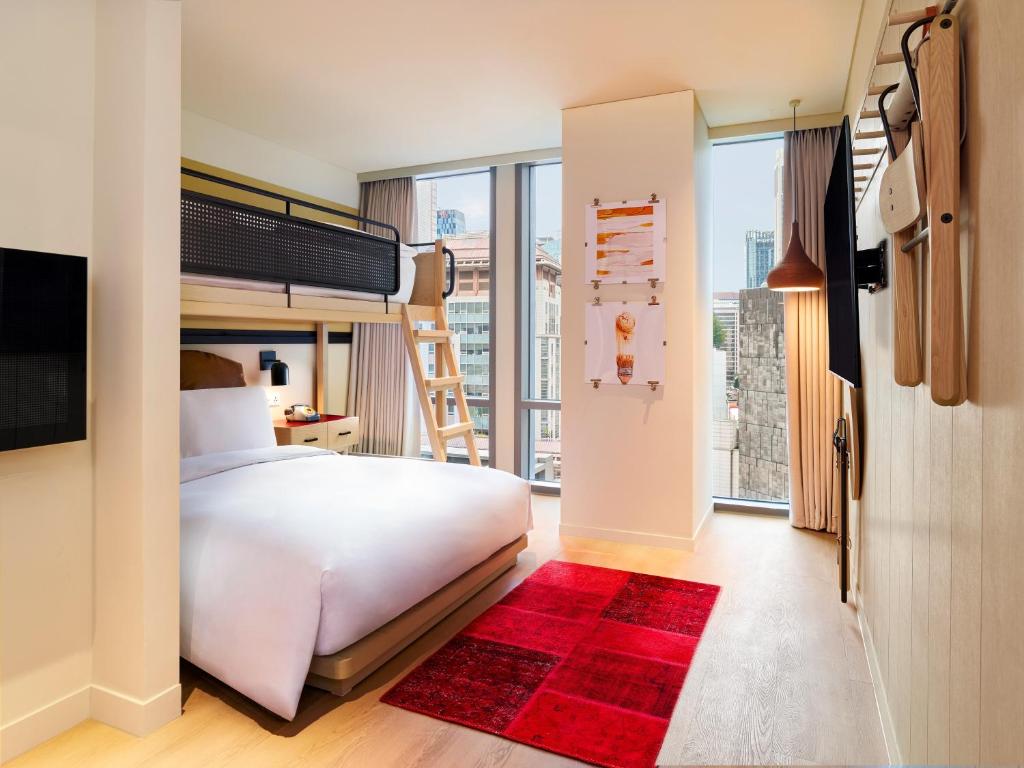 Modern hotel room with a large bed, a lofted bunk bed above it, a wooden ladder, floor-to-ceiling windows with city views, a red area rug, wall-mounted lights, and minimalist décor.