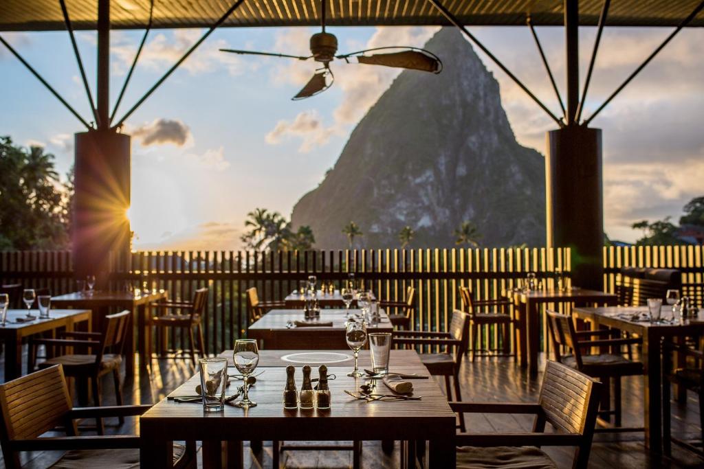 An open-air restaurant with wooden tables set for dining overlooks a dramatic mountain peak at sunset, with warm sunlight streaming in and a ceiling fan above.