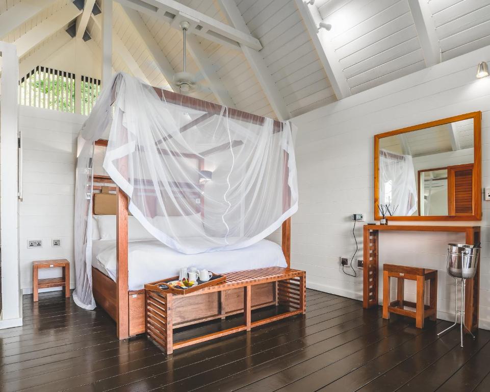 Spacious, airy bedroom with a four-poster bed draped in a white mosquito net, wooden floors, white walls, a wooden bench, a vanity with a mirror, and a breakfast tray on the bed.