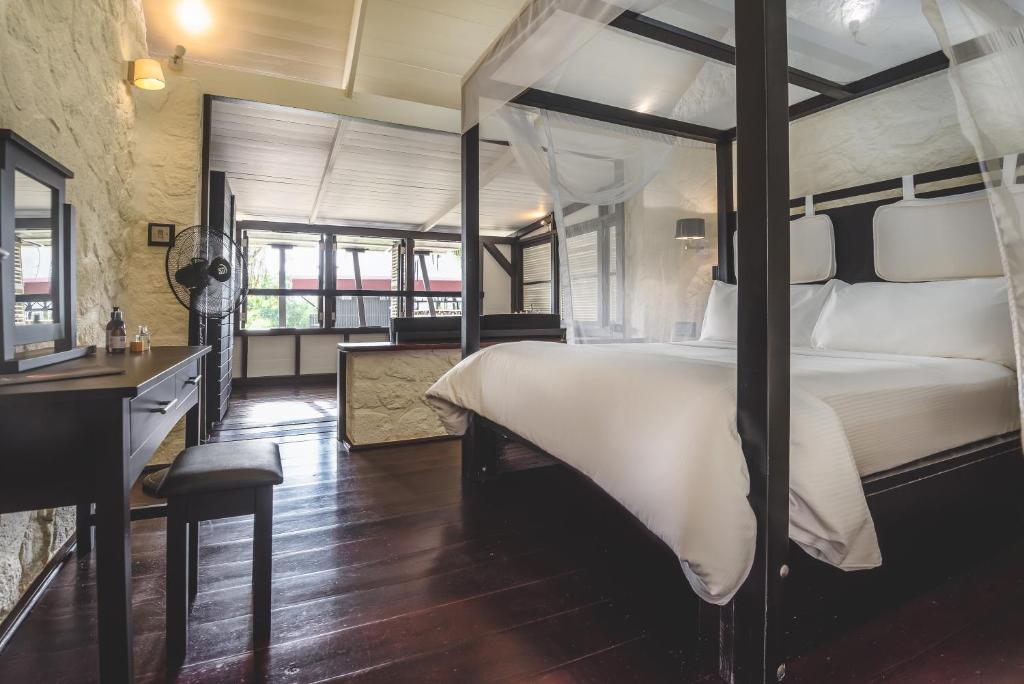 A spacious bedroom with a canopy bed draped in white fabric, dark wooden floors, a dressing table with a stool, a standing fan, and large windows letting in natural light.