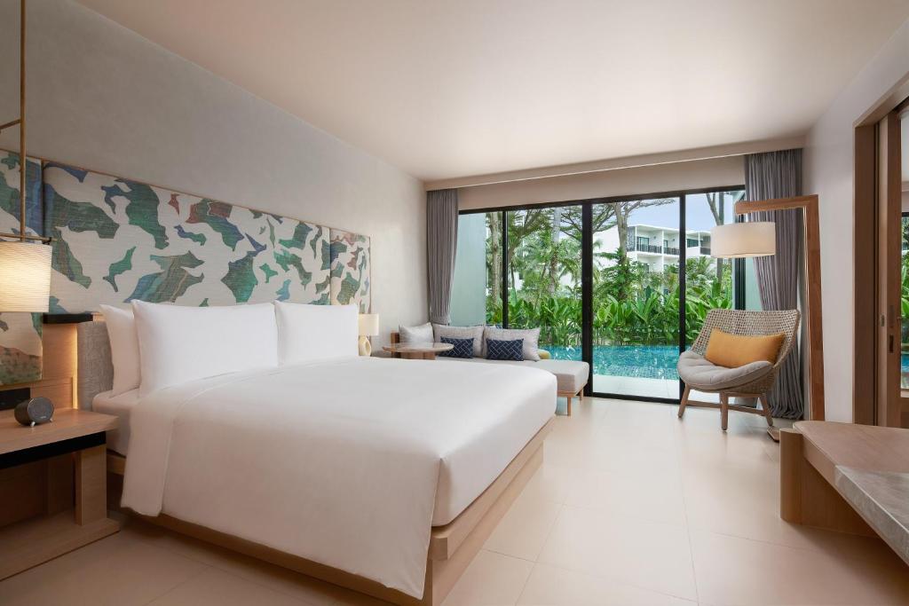 A modern hotel room with a large bed, a colorful headboard, a lounge chair, and a floor-to-ceiling window overlooking a pool and lush greenery outside. The decor is minimal and bright.