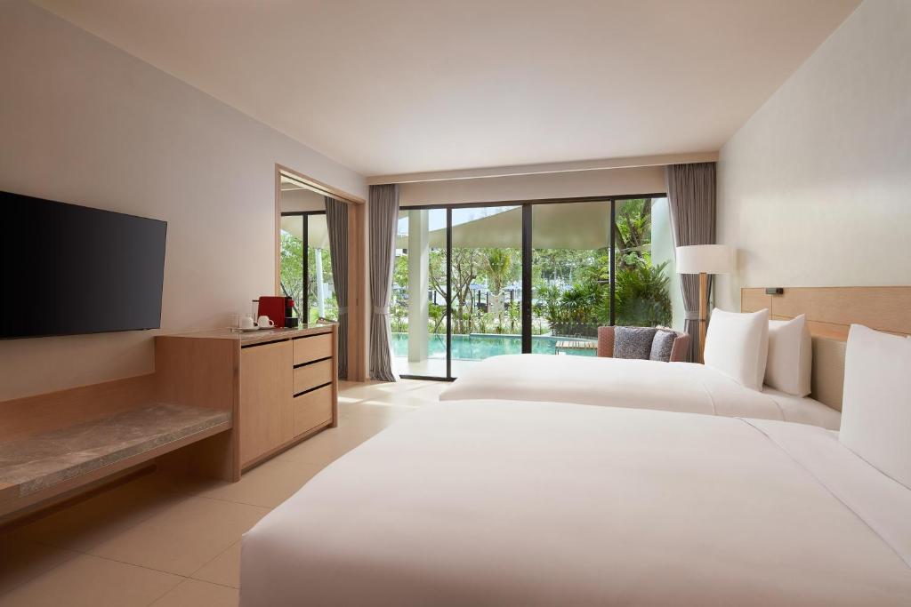 A modern hotel room with two double beds, a wall-mounted TV, a desk with a red kettle, and large glass doors opening to a patio with a view of a pool and lush greenery outside.