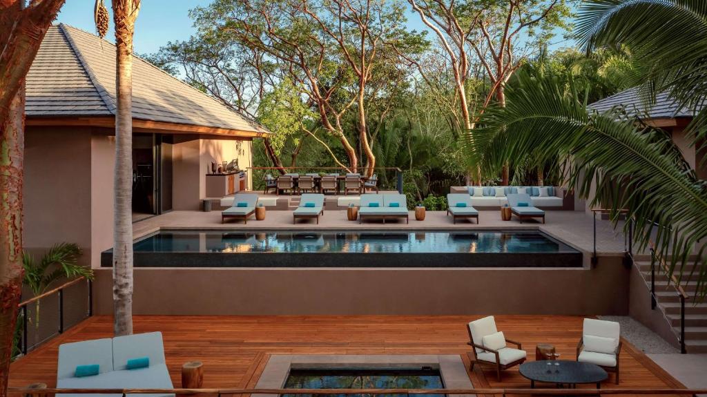 Luxury outdoor pool area surrounded by trees, with cushioned lounge chairs, a bar, and dining area. Wooden deck features additional seating, a small hot tub, and a relaxed, tropical atmosphere.