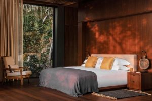 A modern bedroom with wooden walls and flooring, a large bed with white sheets, gray blanket, and gold pillows, a wooden nightstand, an armchair, and a floor-to-ceiling window overlooking lush greenery.