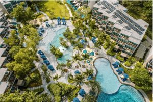 Aerial view of a resort featuring winding pools surrounded by palm trees, sun loungers, and blue umbrellas. Multiple modern buildings with balconies overlook the lush, landscaped pool area.