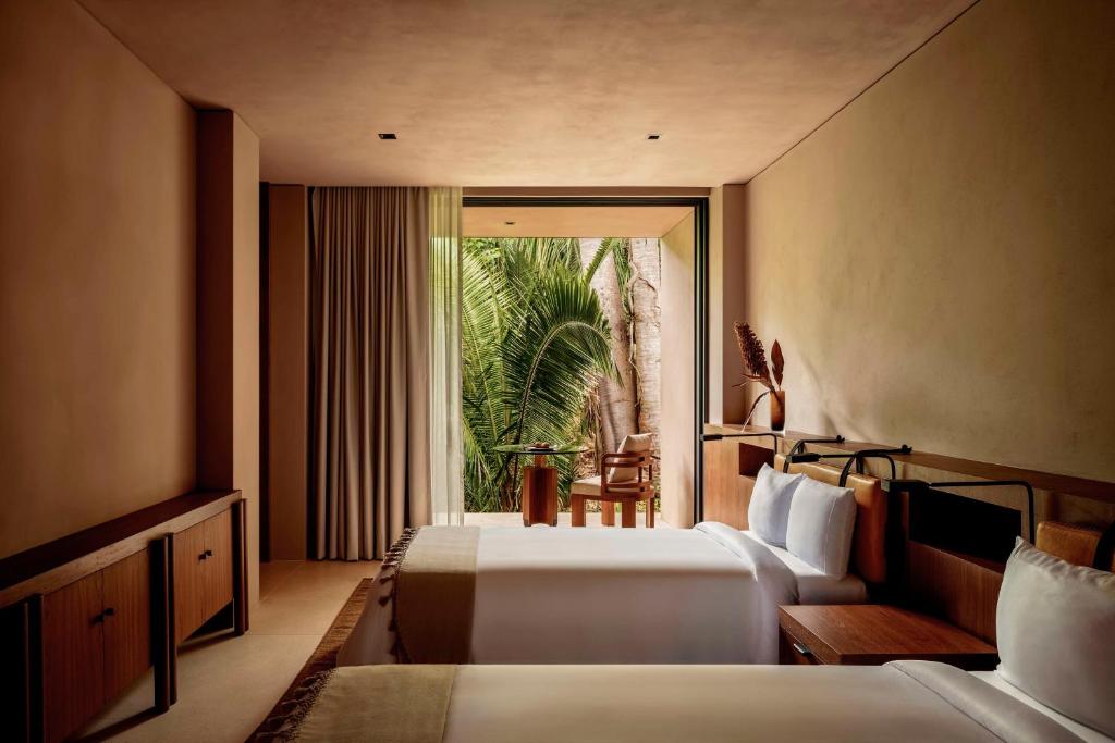 A modern hotel room with two neatly made beds, wooden furniture, soft beige walls, and a large window with curtains, offering a view of lush green palm trees outside.