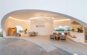 Open, modern lounge area with curved white ceiling, circular hanging lights, wooden furniture, and indoor plants. Veranda sign is on the back wall, and a pool is visible through the open side of the space.