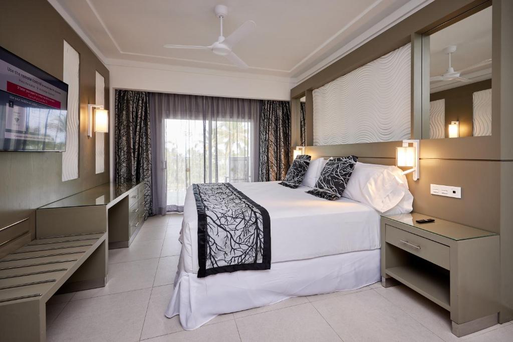 Modern hotel room with a large bed, decorative pillows, bedside lamps, a wall-mounted TV, desk, bench, and large window with curtains offering a view of palm trees outside.