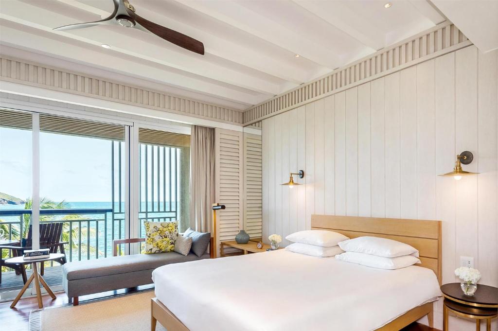 A bright, modern hotel room with a large bed, a lounge chair, wall lamps, and sliding doors leading to a balcony with ocean views. Light wood and white decor create an airy, inviting atmosphere.