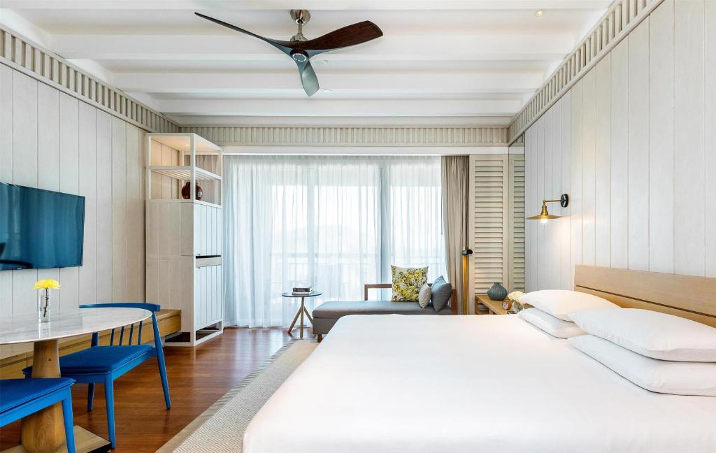 Bright, modern hotel room with a large bed, wall-mounted TV, blue chairs, small table, and a lounge area by floor-to-ceiling windows covered with sheer white curtains. Ceiling fan and wood-accented decor complete the space.