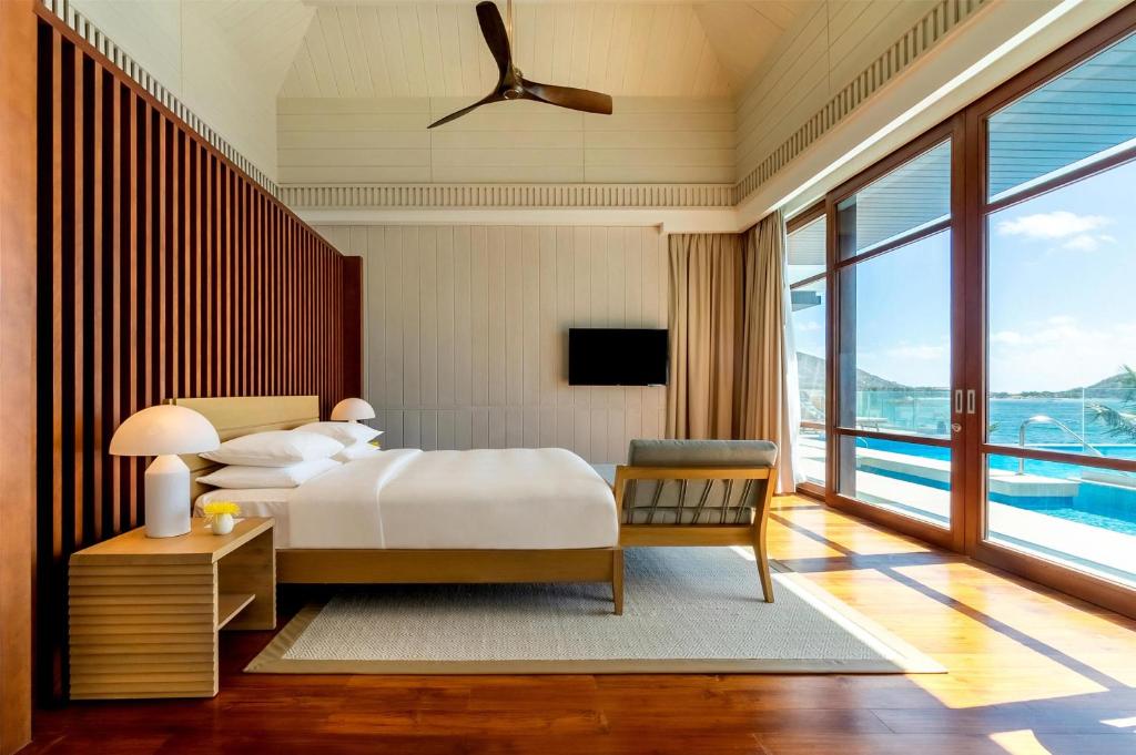 A modern, sunlit bedroom with a large bed, wooden floors, side tables with lamps, a TV, floor-to-ceiling windows, and a view of a private pool and blue sky outside.