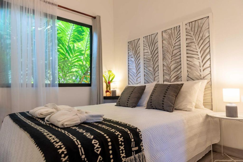 A cozy bedroom with a double bed, white bedding, and black patterned throw, two gray pillows, bedside tables with lamps, a large window with sheer curtains, and leafy plants visible outside. Modern botanical wall art decorates the headboard.