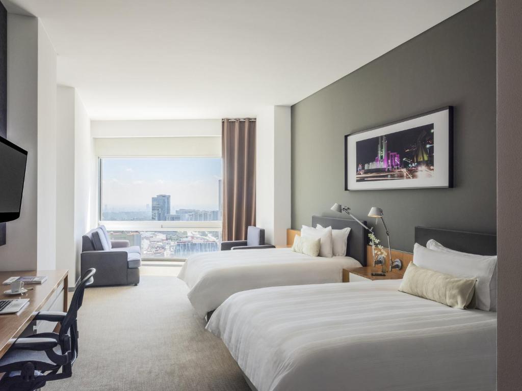 Modern hotel room with two double beds, a large window with city view, a desk with a chair, a gray armchair by the window, and a framed photograph of a cityscape above the beds.