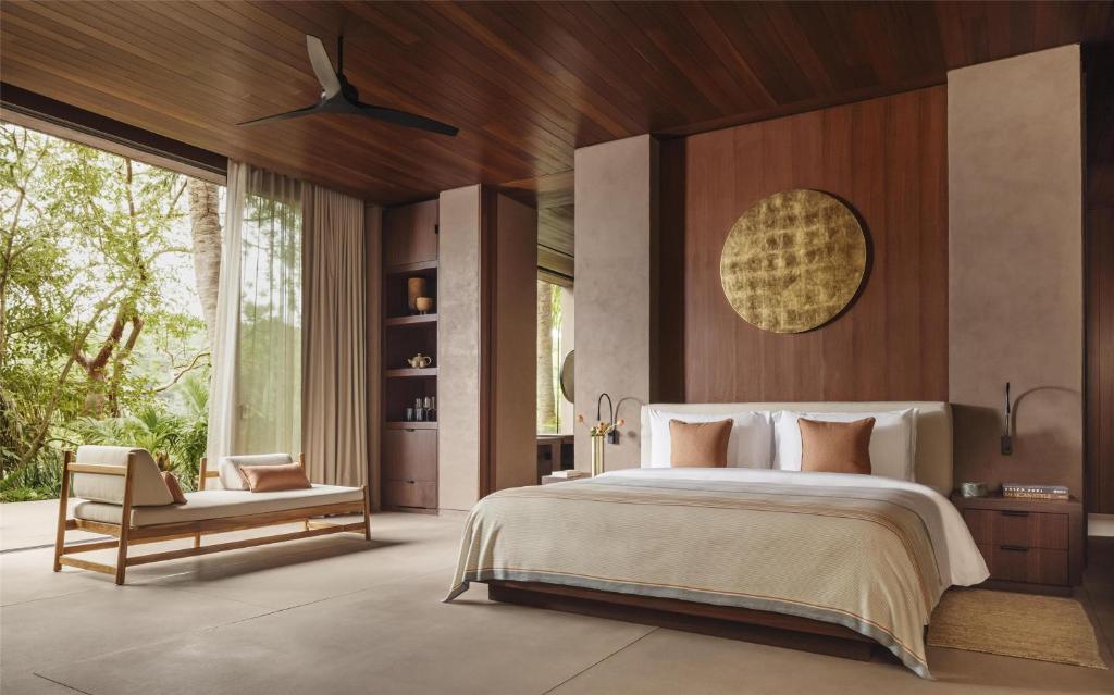 Modern bedroom with wood accents, a large bed with white and brown bedding, a lounge chair, floor-to-ceiling windows revealing greenery, and a gold circular wall art above the bed. Natural light fills the room.