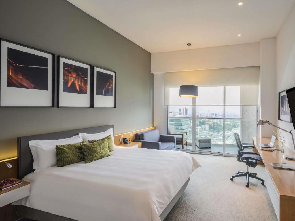 Modern hotel room with a large bed, green accent pillows, wall art above the headboard, a sofa, desk with chair, and floor-to-ceiling window overlooking a city skyline with a balcony. Room is bright and tidy.