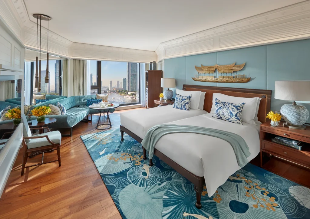 Luxurious hotel room with two beds, blue and white decor, floral rug, seating area by large windows, city skyline view, and elegant wall art above the headboard.