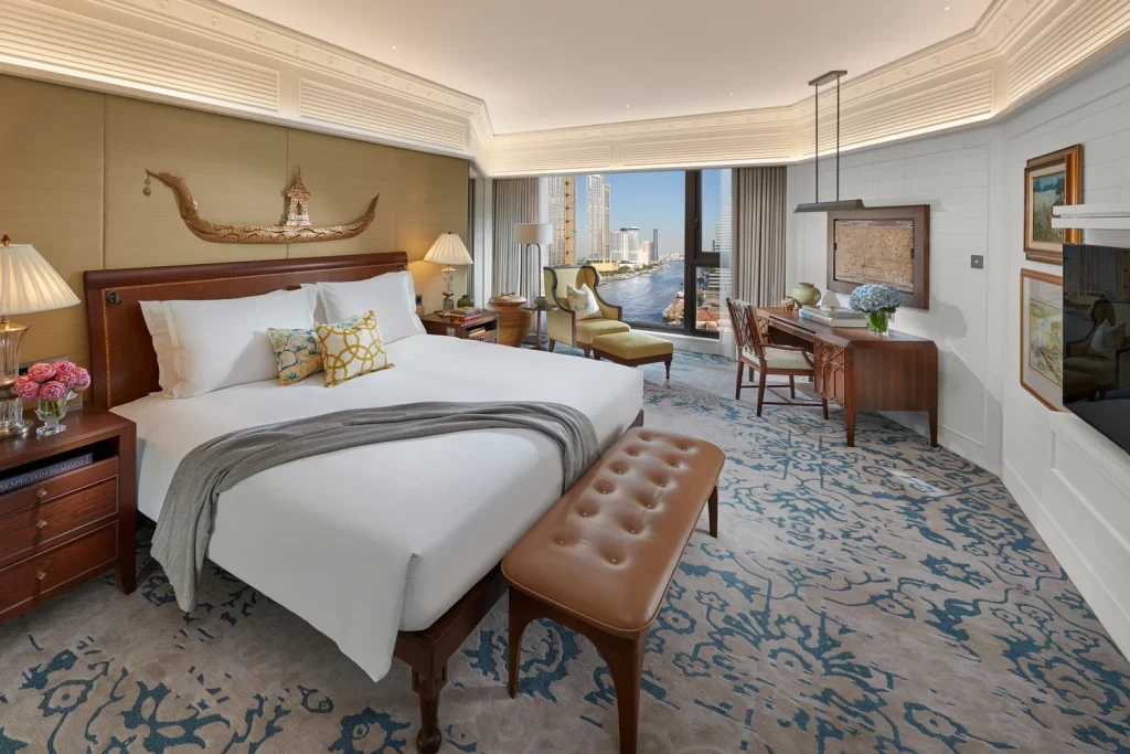 Luxurious hotel room with a large bed, patterned carpet, elegant decor, desk, seating area, large windows, and a city and river view. Warm lighting and decorative accents create a welcoming atmosphere.