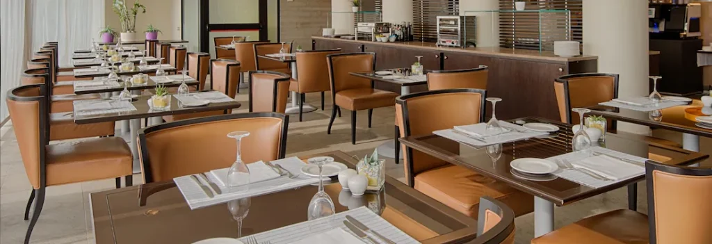 A modern restaurant dining area with neatly set tables, brown chairs, glassware, and tableware. The space is well-lit with natural light and features potted plants and a buffet counter in the background.
