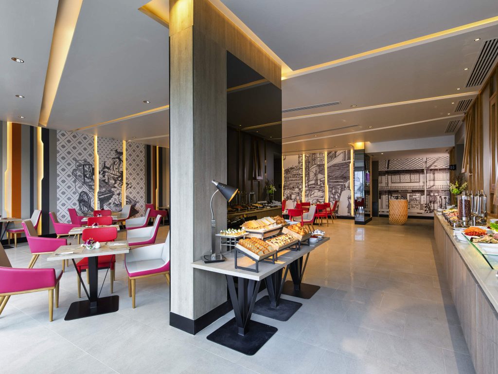 Modern hotel dining area with pink and white chairs, wooden tables, and a buffet setup featuring pastries and breakfast items. Walls have artistic patterns and black-and-white mural artwork. Bright, welcoming atmosphere.