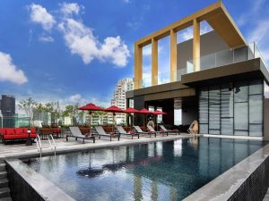Rooftop pool with lounge chairs and red umbrellas, modern building in the background, and city skyline under a bright blue sky with scattered clouds.