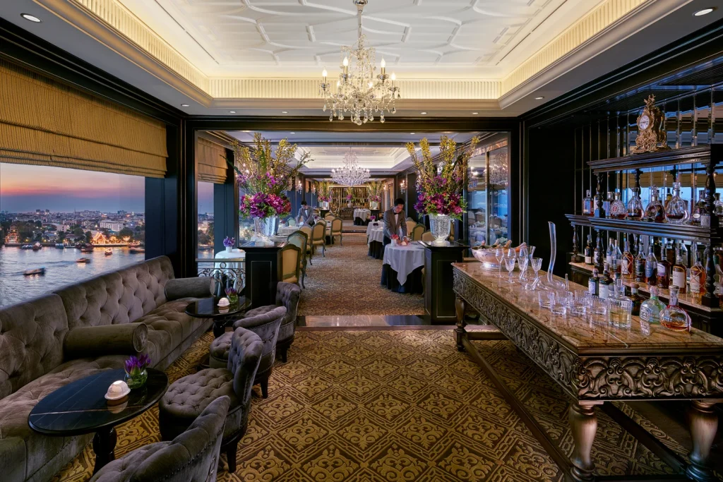 Elegant restaurant interior with chandeliers, plush seating, ornate carpets, and a well-stocked bar. Large windows offer a sunset city and river view, while tables are set with white linens and floral arrangements.
