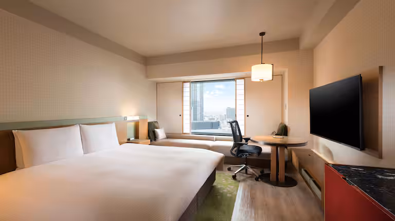 A modern hotel room with a large bed, a wall-mounted TV, a desk and chair, a lounge seat by the window, and a city view visible through the window. The decor is minimalist and neutral-toned.