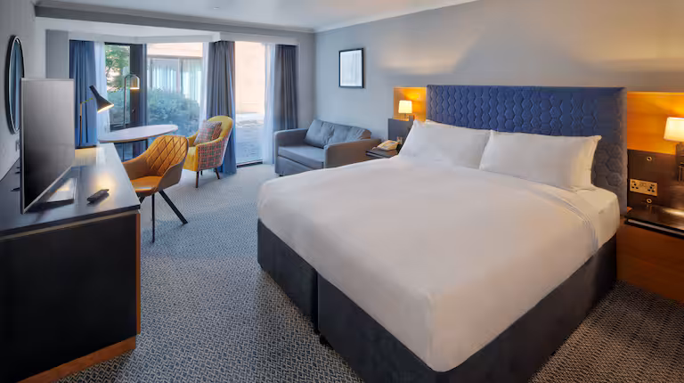 Modern hotel room with a large bed, blue headboard, sofa, desk with two yellow chairs, TV, and floor-to-ceiling windows with curtains letting in natural light.