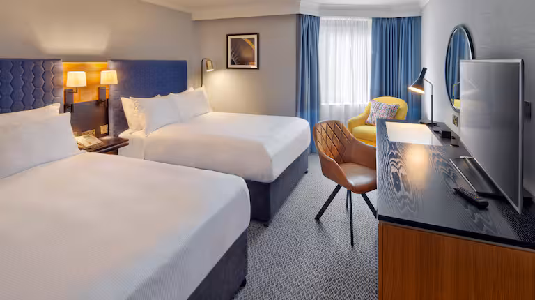 Modern hotel room with two large beds, a desk with a chair, a TV, and a yellow armchair near a window with blue curtains. Walls are light gray, and lighting is soft and inviting.