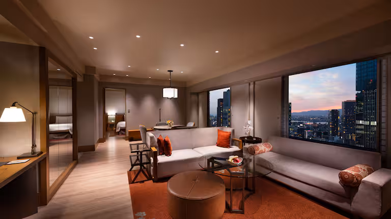 A modern hotel suite with a spacious living area, plush sofas, a desk, and large windows showing a city skyline at sunset. Warm lighting and neutral tones create a cozy, elegant atmosphere.