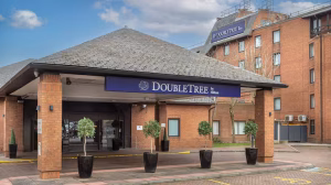 The entrance of a DoubleTree by Hilton hotel, featuring a covered driveway, potted plants, and a brick building with the DoubleTree logo visible on both the canopy and the side of the building.