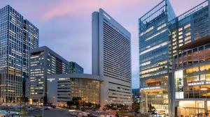 A cityscape at dusk featuring tall, modern glass and concrete office buildings with lights on inside, and blurred traffic in the foreground. The sky is tinged with soft purple and pink hues.