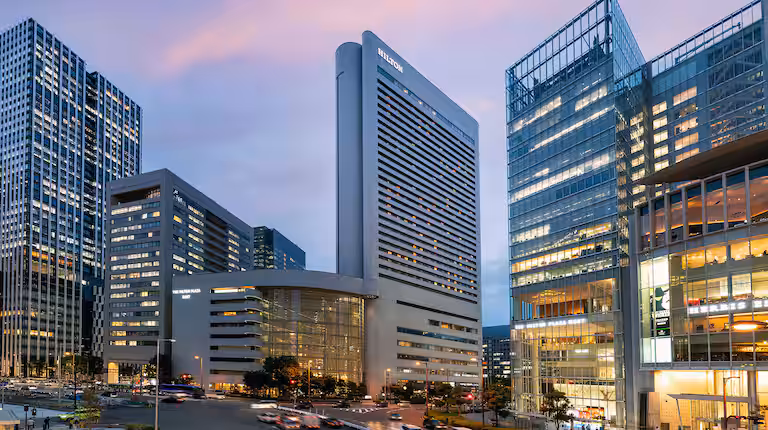 A cityscape at dusk featuring tall, modern glass and concrete office buildings with lights on inside, and blurred traffic in the foreground. The sky is tinged with soft purple and pink hues.
