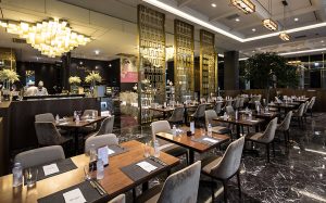 Modern restaurant interior with neatly arranged tables and chairs, elegant chandeliers, dark marble flooring, gold room dividers, and decorative plants, creating a luxurious and inviting dining atmosphere.