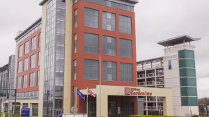 A modern multi-story Hilton Garden Inn hotel with large windows and red brick facade, two flags flying outside, and a multi-level parking structure in the background on a cloudy day.