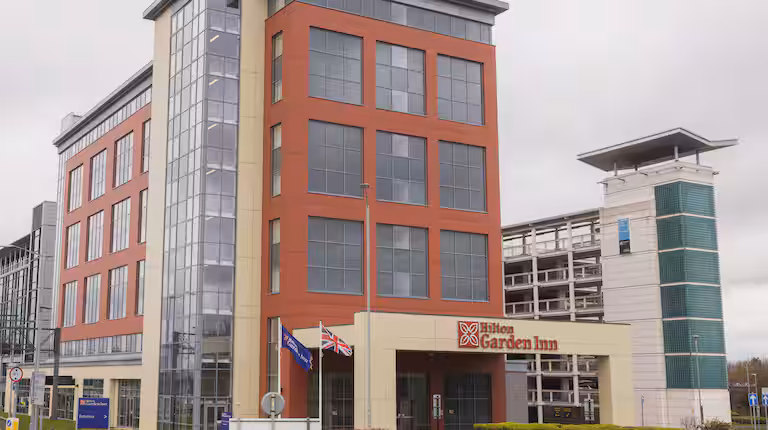 A modern multi-story Hilton Garden Inn hotel with large windows and red brick facade, two flags flying outside, and a multi-level parking structure in the background on a cloudy day.