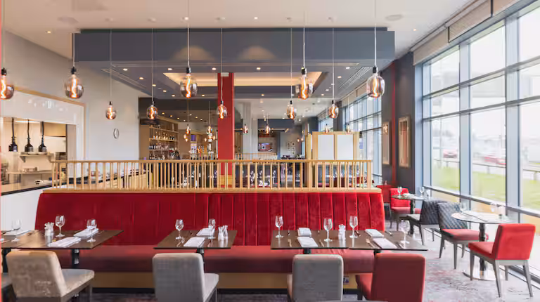 Modern restaurant interior with large windows, red upholstered booth seating, tables set with glasses and cutlery, hanging pendant lights, and a view of an open kitchen area in the background.