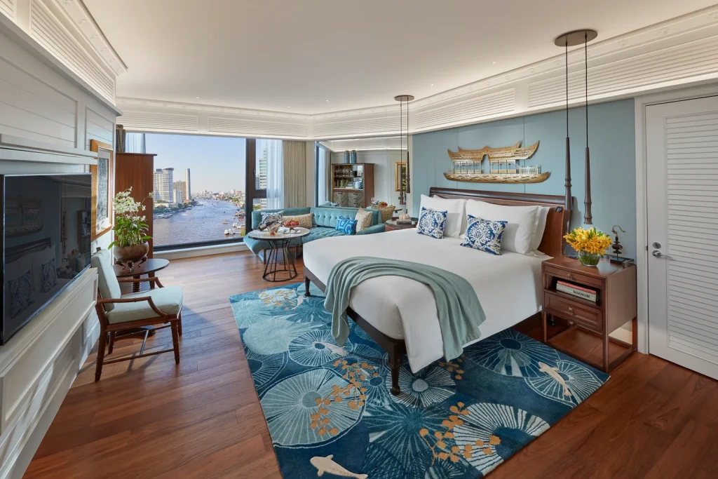 A spacious hotel room with a large bed, blue-themed decor, a city and river view through wide windows, a cozy seating area, flowers, and elegant wooden furniture on a patterned blue rug.