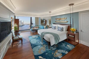 A spacious hotel room with a large bed, blue-themed decor, a city and river view through wide windows, a cozy seating area, flowers, and elegant wooden furniture on a patterned blue rug.