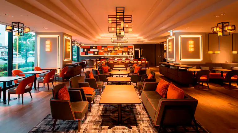 A modern lounge area with cozy seating, orange and brown chairs, sofas, wooden tables, warm lighting, and a stylish bar at the back, creating a welcoming atmosphere. Large windows let in natural light.
