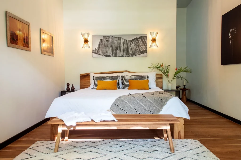 A modern bedroom with a large bed featuring white bedding, striped and yellow pillows, a wooden bench at the foot, wall-mounted nightstands, art on the walls, and a patterned rug on a wooden floor.